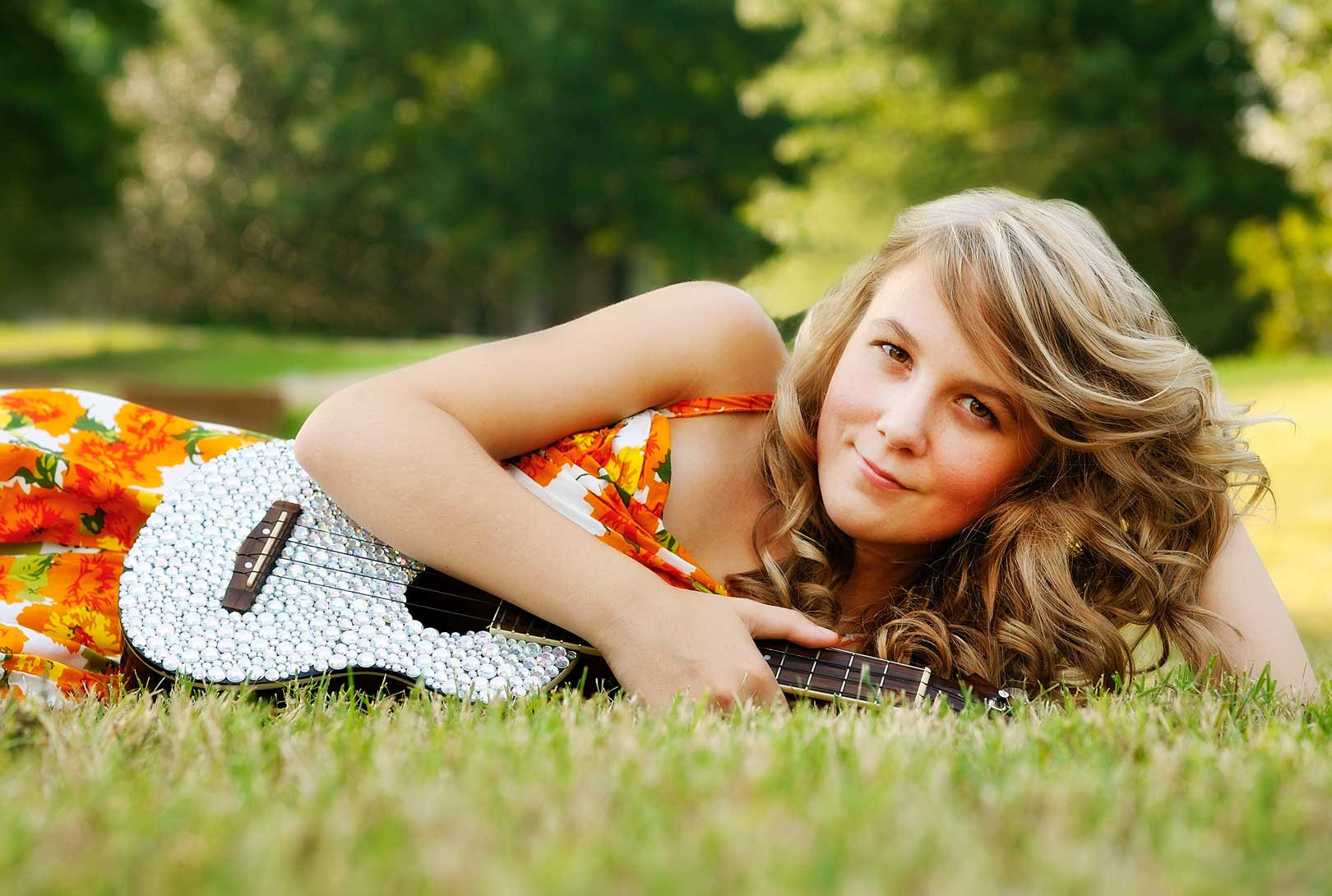 Carlie Mahnke lying in grass holding sparkly ukulele wearing an orange white and red floral dress