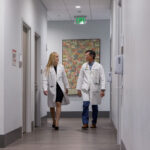 A male and a female doctor wearing lab coats walking down a hallway at a healthcare facility