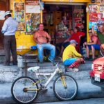 men smiling sitting outside a store with yellow walls a bike and red car parked in San Vicente