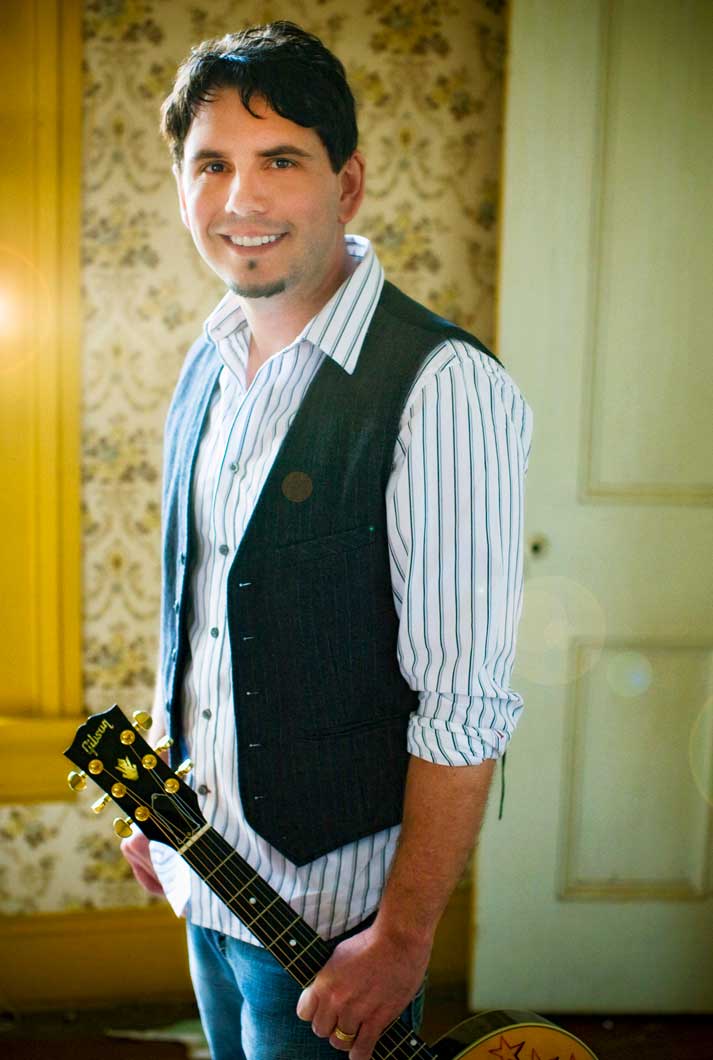 Joe Zelek in striped white shirt and black vest in home with floral wallpaper holding guitar