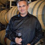 Kip Summers wearing black dress shirt holding glass of red wine standing near wine barrels at winery