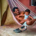 man holding child while lying in hammock hanging on front yellow and red porch in Bahia de Caraquez