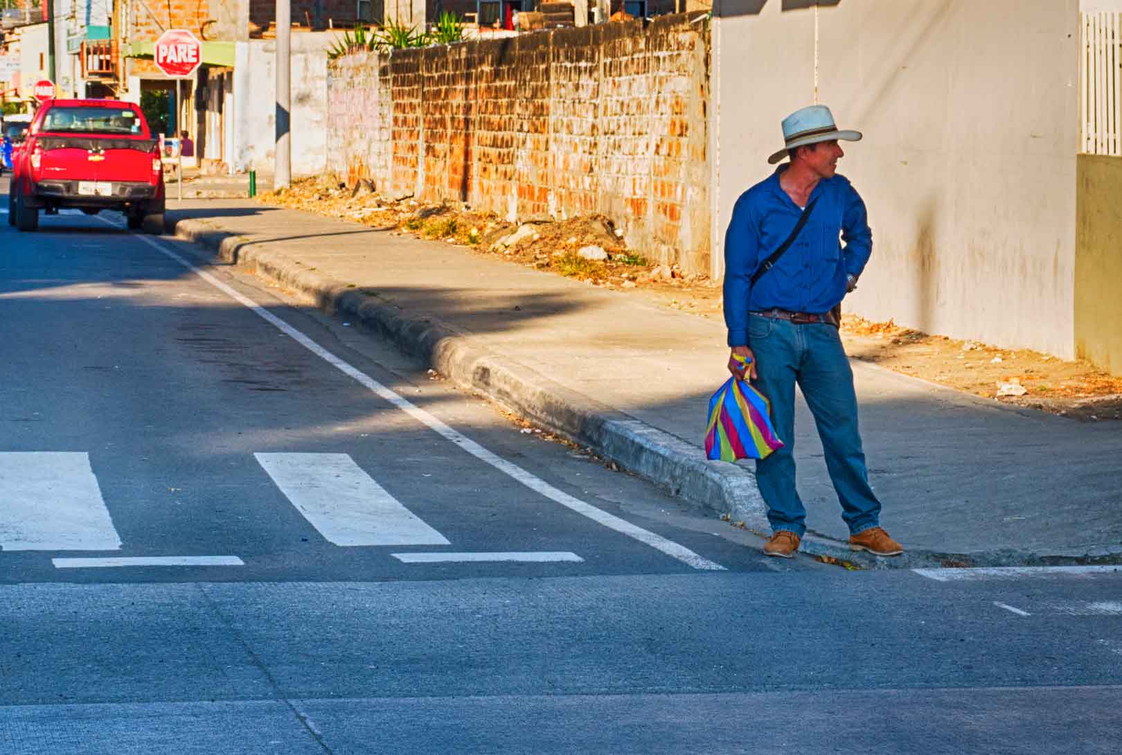man standing on street corner wearing jeans a blue shirt and Panama hat holding colorful bag Ecuador
