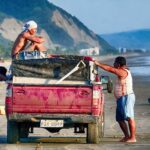 Three men in shorts with red work truck one with head wrapped in shirt on Canoa beach in Ecuador