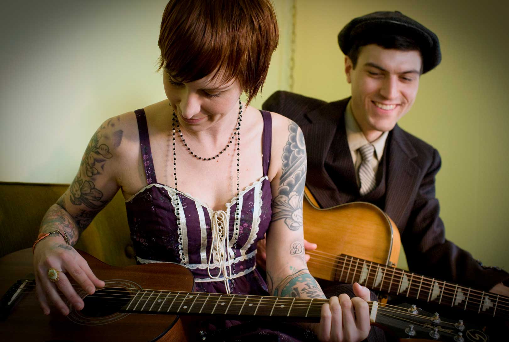 Kate with tattooed arms wearing purple dress with Steven in vintage suit and hat playing guitars