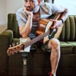 cancer amputee Travis Meadows with his guitar sitting on a green retro couch