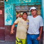 two Ecuadorean men with arms around each other standing in front of blue and purple building smiling