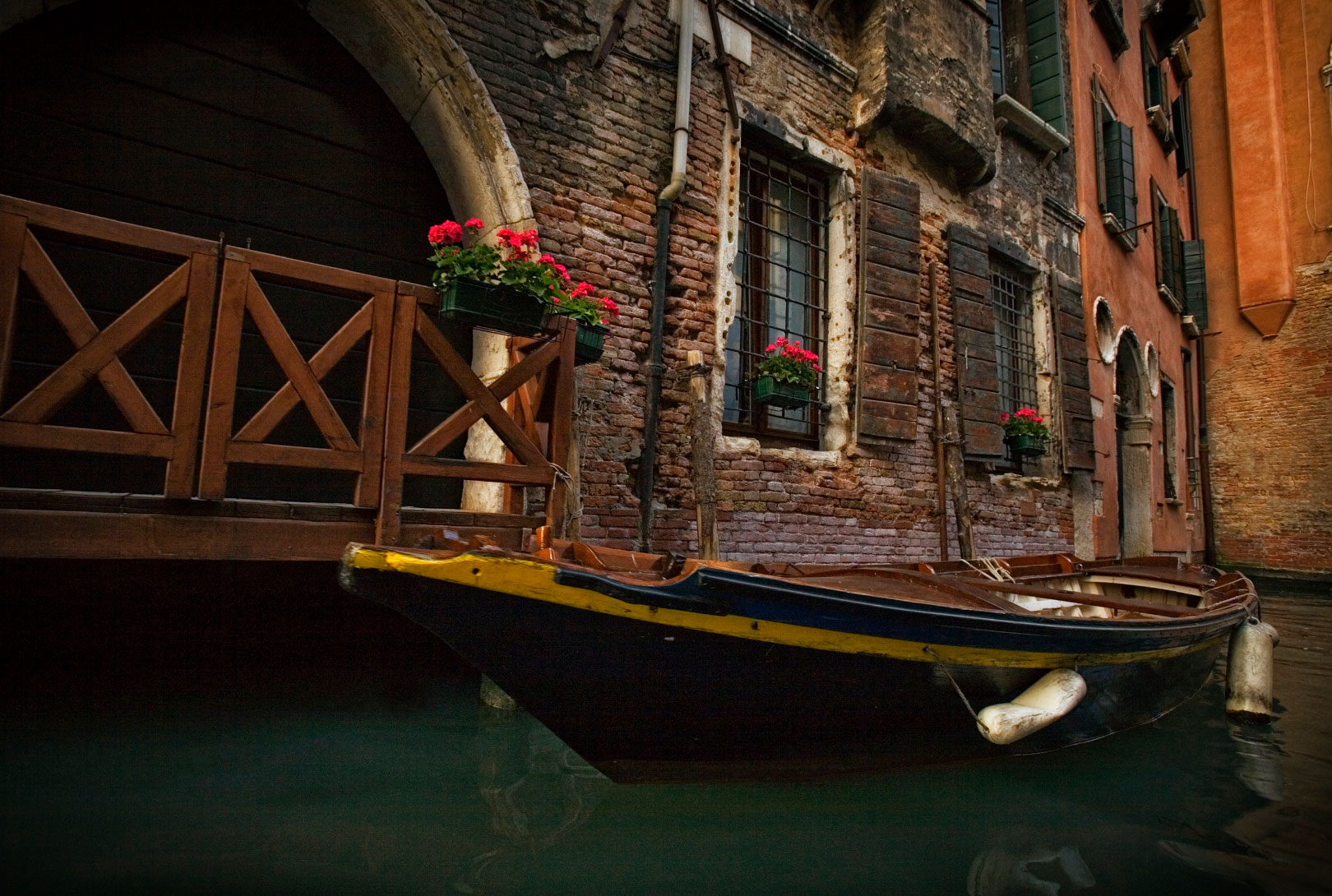 wooden boat with yellow trim tied to a brick building with red flowers in canal Venice Italy