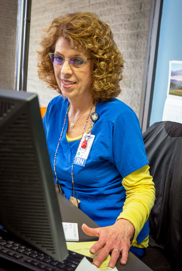 female nurse wearing blue scrubs and glasses sitting at her computer working in her office