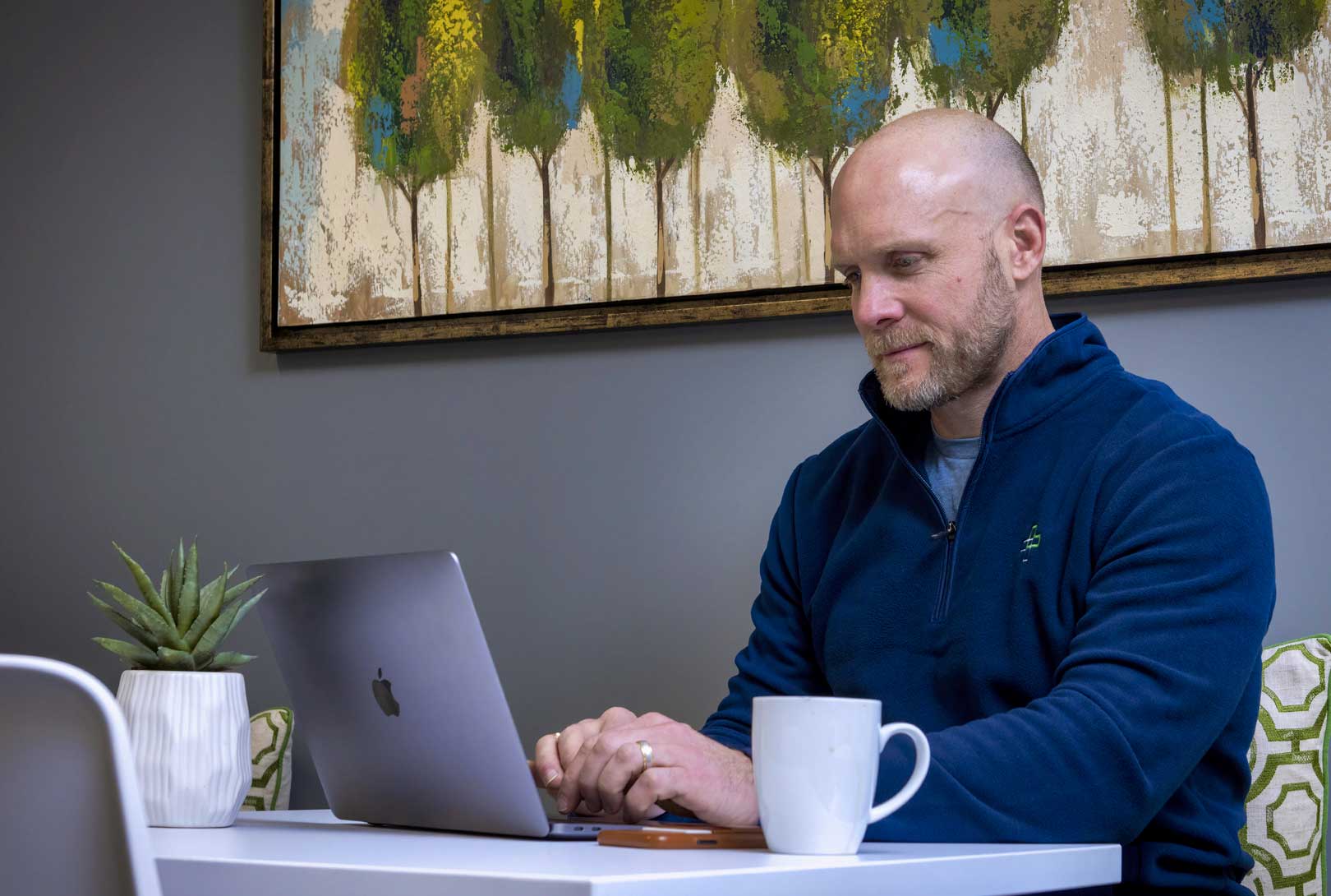 Bo Bartholomew in blue pullover sitting at white table working on his laptop with painting of trees in background