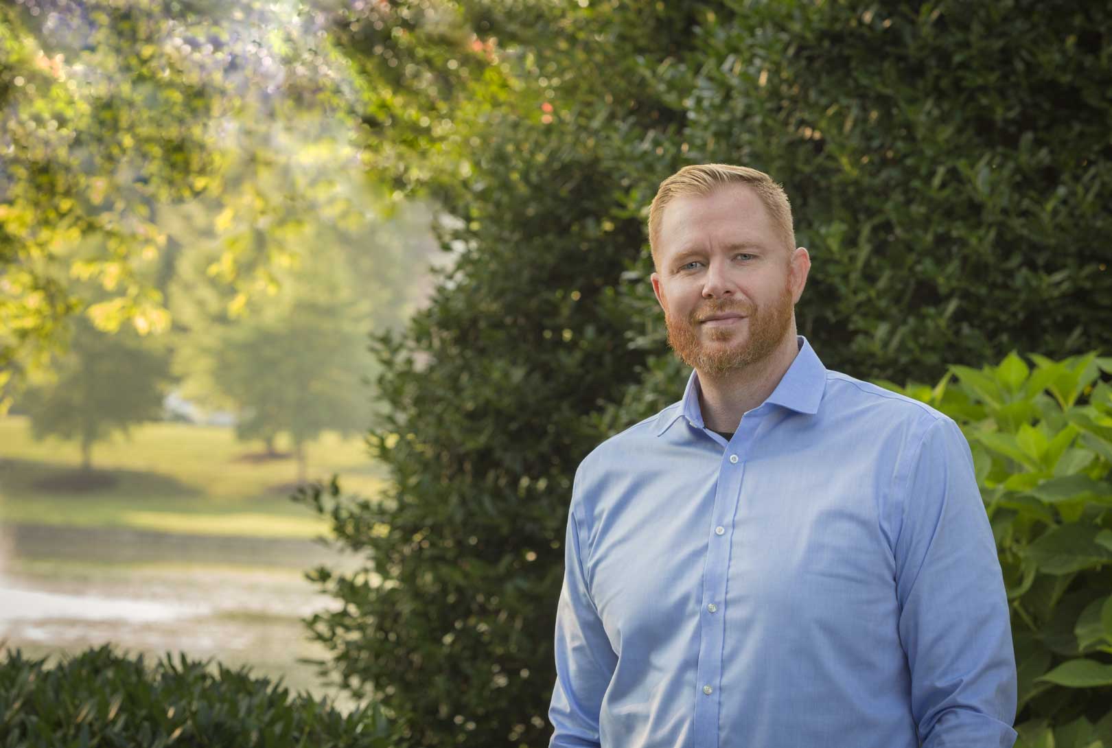 executive headshot of David Dutton in blue button up shirt with trees and park in background
