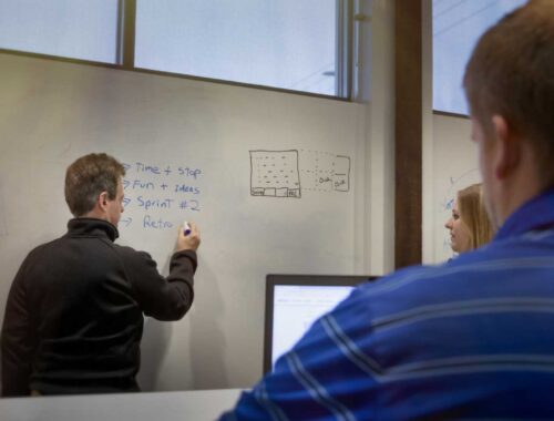 a white man in black pullover shirt writing on a whiteboard during a meeting at L3Harris