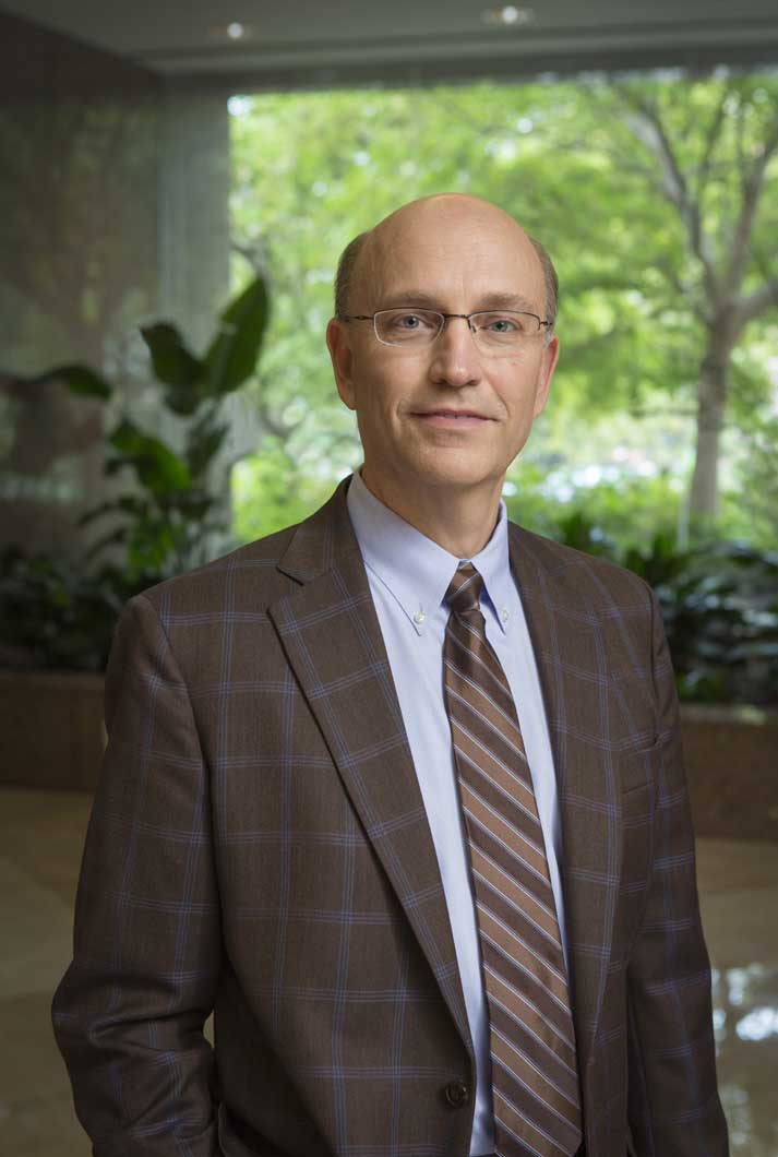 executive headshot of Mark Robbins in brown striped suit, tie, light blue shirt standing in lobby