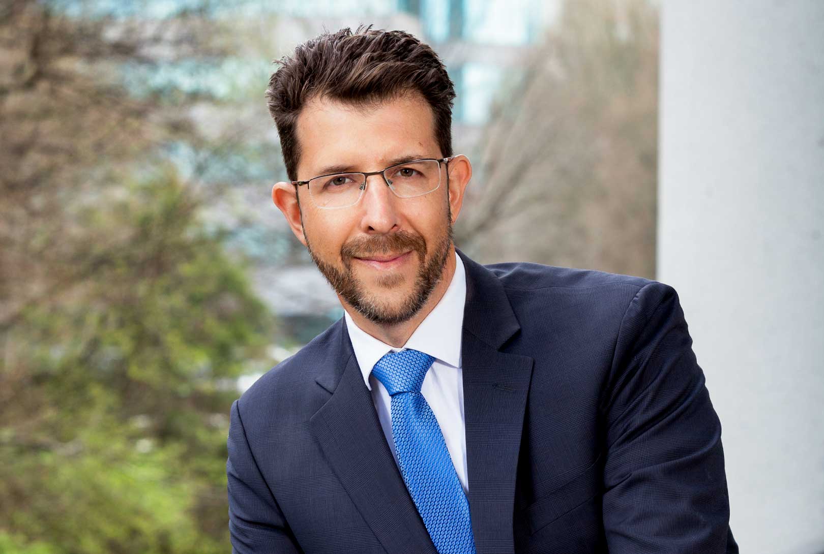 outdoor pr headshot of executive Rene Marzagao wearing a blue suit and tie with glasses