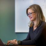 woman executive with glasses in conference room with whiteboard in background working on laptop