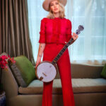 Ashley Campbell standing in front of window with green curtains and couch wearing red holding banjo