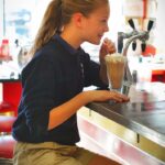 young blonde girl in shorts with a ponytail sitting on a bar stool drinking a milkshake