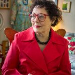 Jane Snyder in her home wearing a red coat and red-rimmed glasses