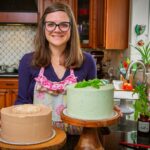 baker Kari Suh standing at her kitchen counter with two beautiful cakes