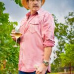 Kix Brooks wearing a pink shirt and white hat in his vineyard with wine
