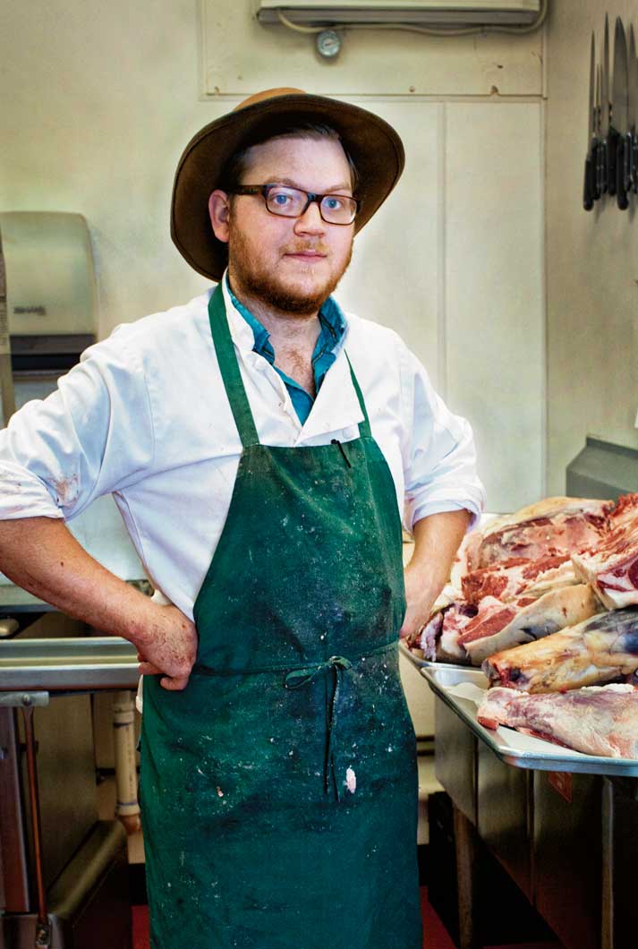 Butcher Tim George standing near his carved raw meat in hat, glasses and green apron