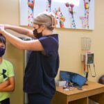 female nurse in dark scrubs measures height of boy in a mask and green shirt in a children's clinic