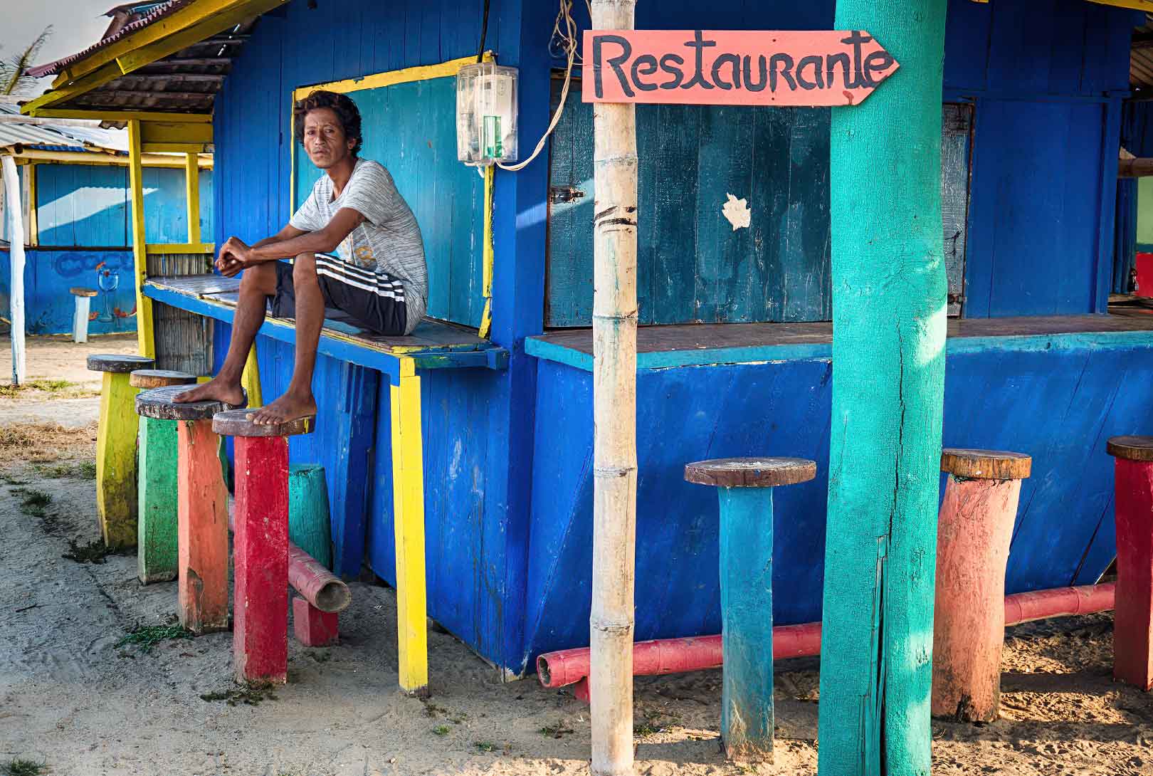 A man sitting on bar with stools painted in bright blue, green, red, orange yellow in Canoa Ecuador