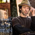 Travis Meadows in a hat sitting in the courtyard smoking a cigarette
