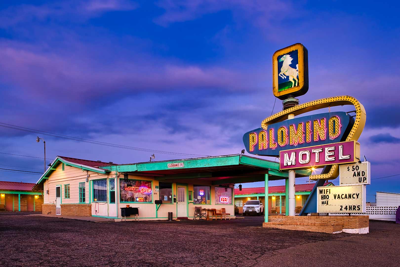 Vintage-style motel with neon signs, featuring "Palomino Motel" and room rates. Offers amenities like WiFi, HBO, and 24-hour service. Evening sky in the background.