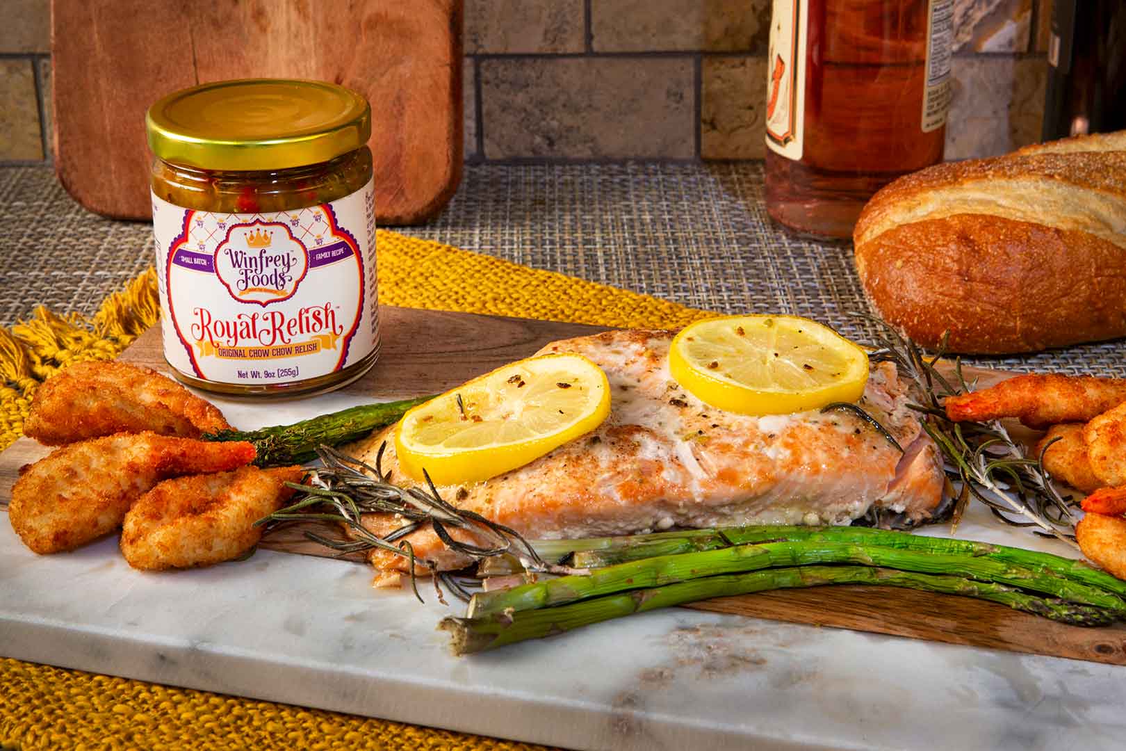Grilled salmon topped with lemon slices on a board with asparagus, shrimp, bread, and a jar of Royal Relish.