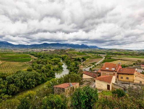 Travel photography of a rural landscape featuring a river, vineyards, and fields, with houses in the foreground and mountains under a cloudy sky in the distance in Briones, Spain.