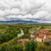 Travel photography of a rural landscape featuring a river, vineyards, and fields, with houses in the foreground and mountains under a cloudy sky in the distance in Briones, Spain.
