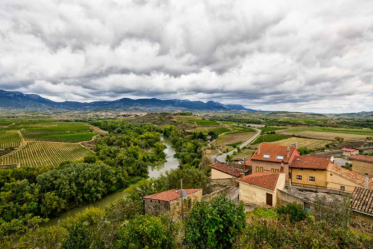 Travel photography of a rural landscape featuring a river, vineyards, and fields, with houses in the foreground and mountains under a cloudy sky in the distance in Briones, Spain.