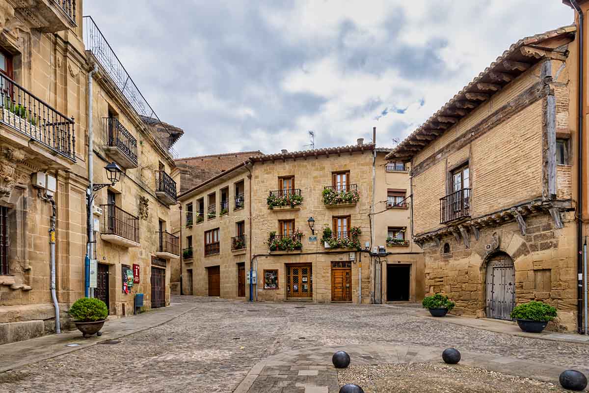 A quiet, cobblestone town square bordered by historic stone buildings with wooden balconies and flower boxes, under a cloudy sky in Briones, Spain.