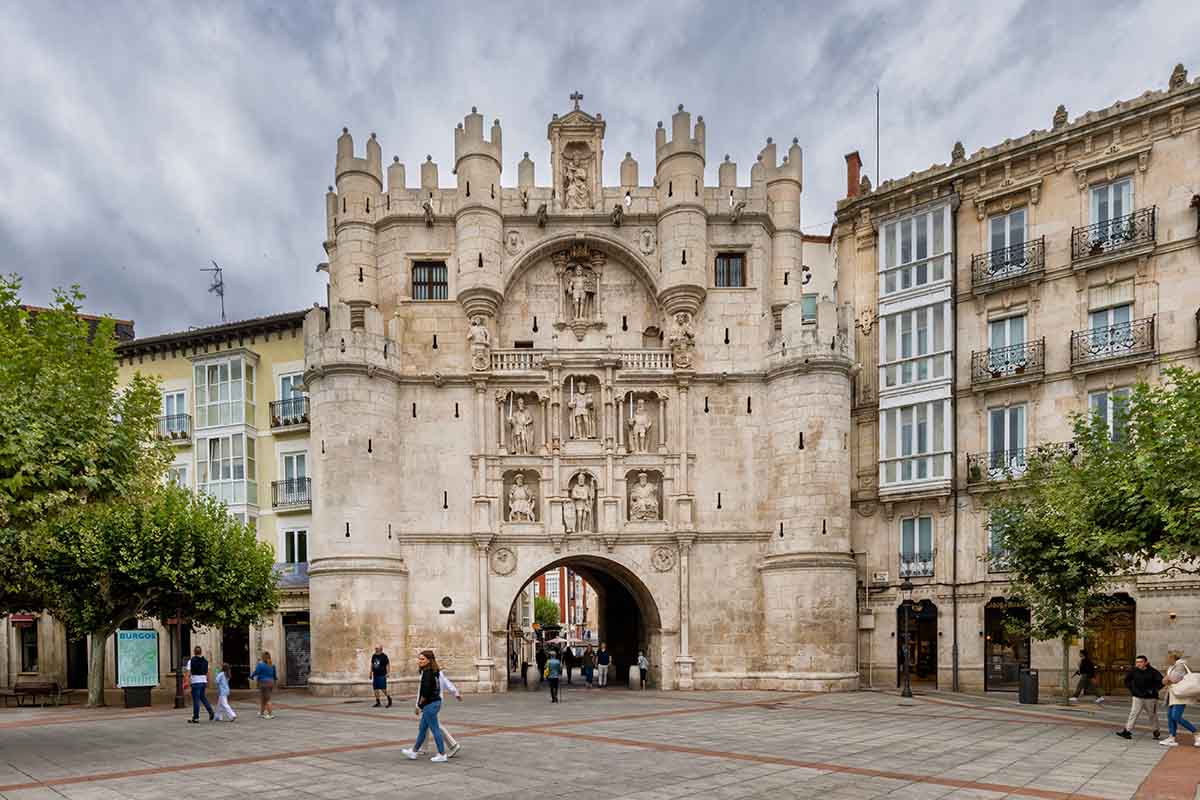 The large medieval stone gate with intricate carvings and statues stands in a public square, surrounded by historic buildings and people walking nearby in Burgos, Spain.