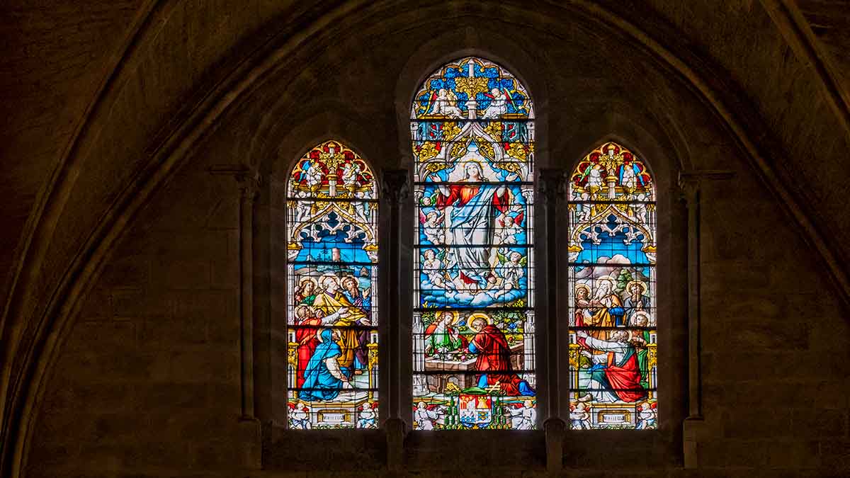 Stained glass window with three arched panels depicting religious scenes, set in a stone wall inside the Burgos Cathedral in Spain.