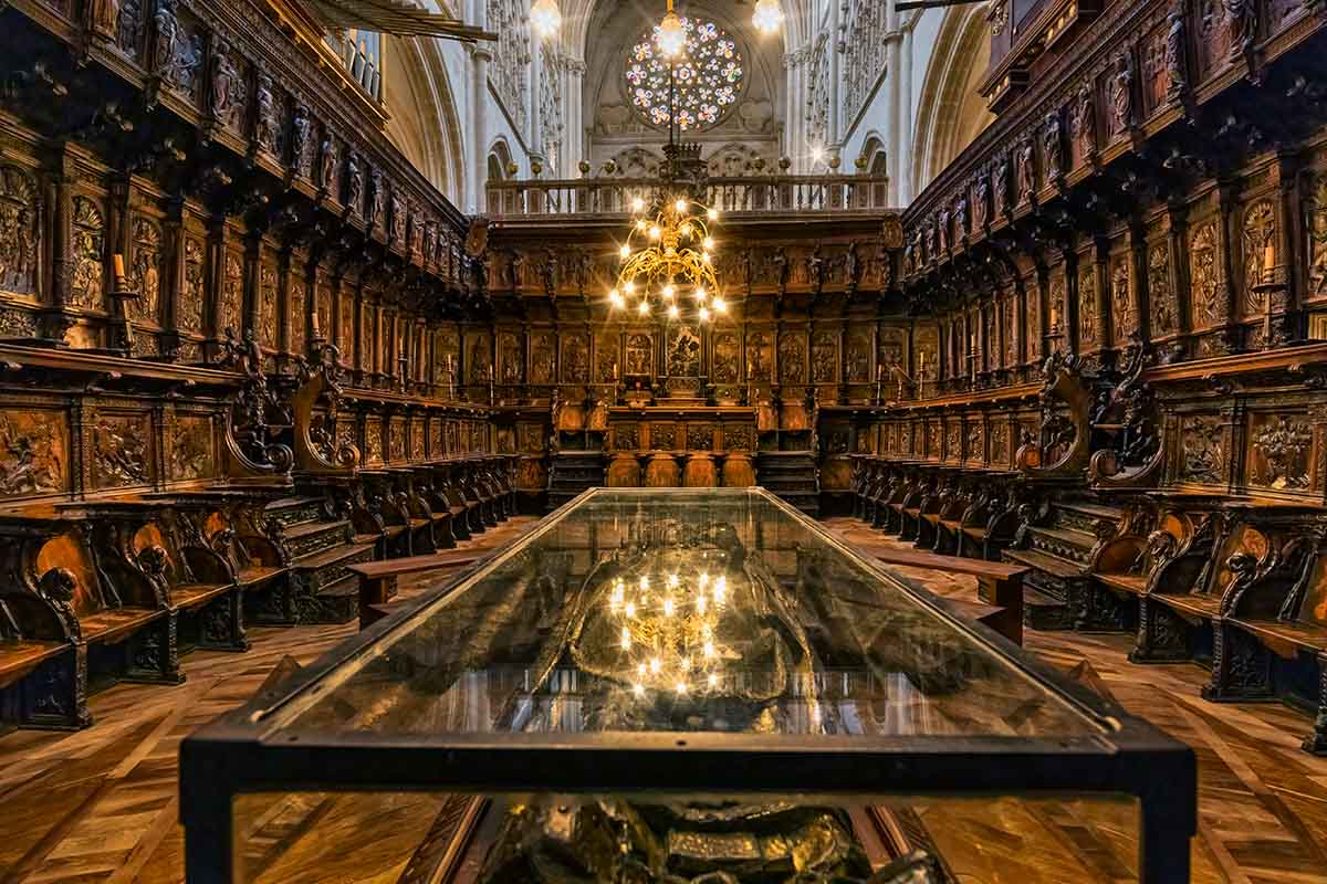 Ornate wood-paneled choir stalls line both sides of the historic Burgos cathedral interior, with a glass-topped table in the center and a chandelier hanging above in Spain.
