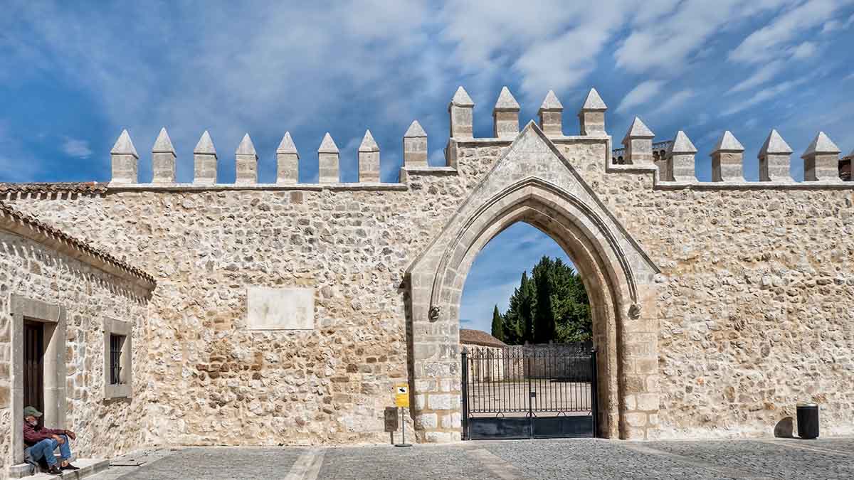 A stone medieval gate with pointed arches and battlements, set within a high wall. A person sits on the left side near a doorway under a partly cloudy sky at the Huegas Monastery in Burgos, Spain.