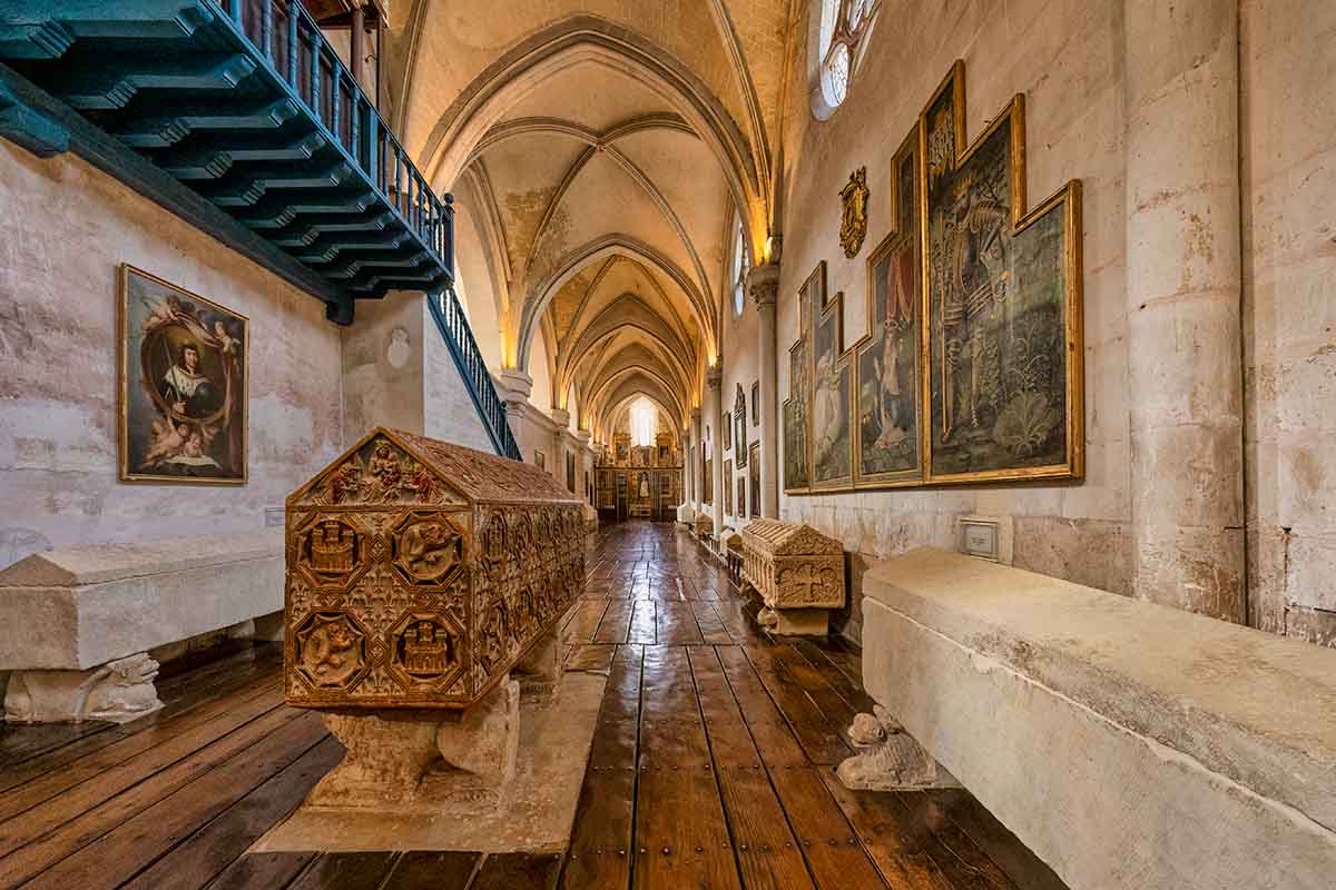 A stone crypt with intricate carvings stands in a vaulted corridor lined with paintings, wooden floors, and benches under arched ceilings in the Huegas Monastery in Burgos, Spain.