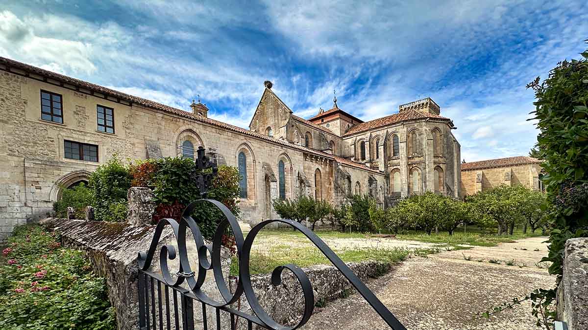 Stone building with arched windows and a gabled roof, surrounded by gardens and an iron gate under a partly cloudy sky at the Huegas Monastery in Burgos, Spain.