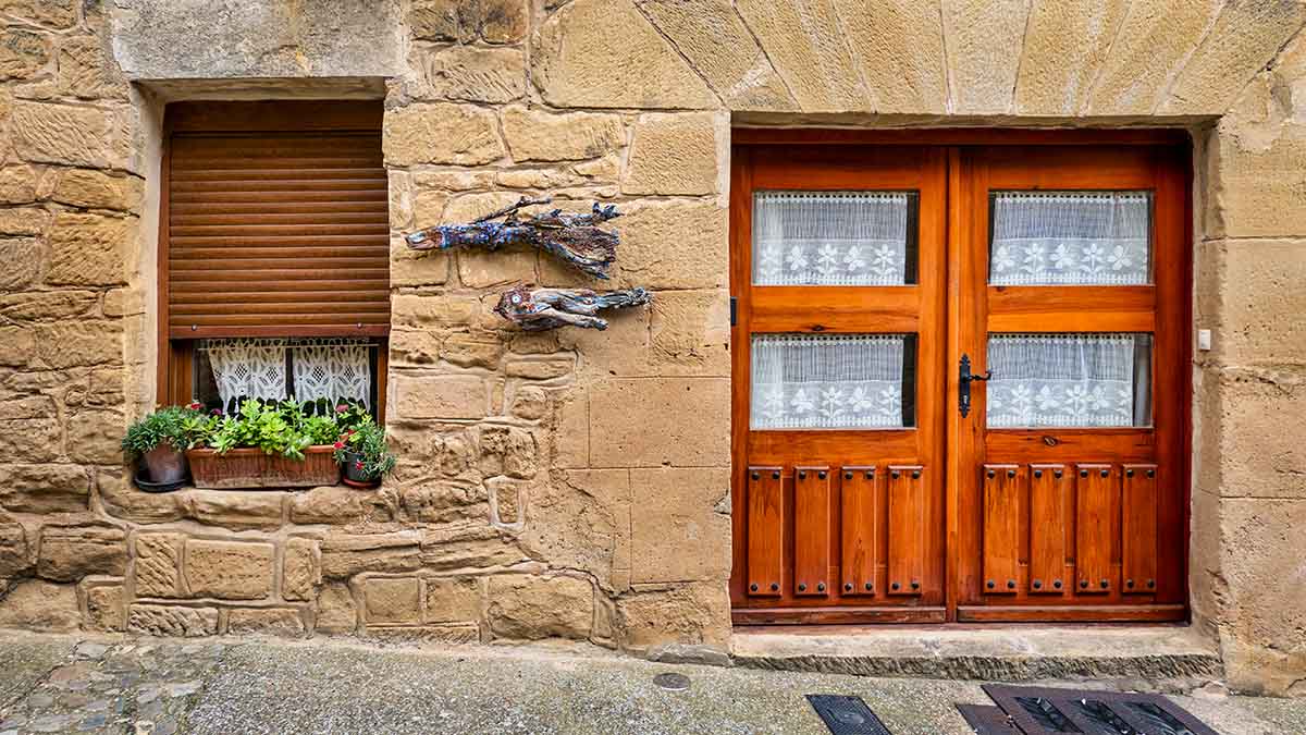 A stone building facade with a closed wooden window shutter, a flower box with green plants, and a wooden double door with lace curtains. Driftwood decor is mounted on the wall in Sajazarra, Spain