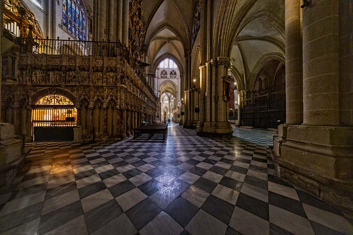 Inside the Toledo cathedral in Spain, with arched ceilings, stone columns, ornate carvings, and a checkered black-and-white tile floor.
