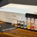 An Epson Ecotank ET-8550 printer sits on a wooden table next to five bottles of Epson ink and a black plastic tray, ready to unleash creativity in any workspace.