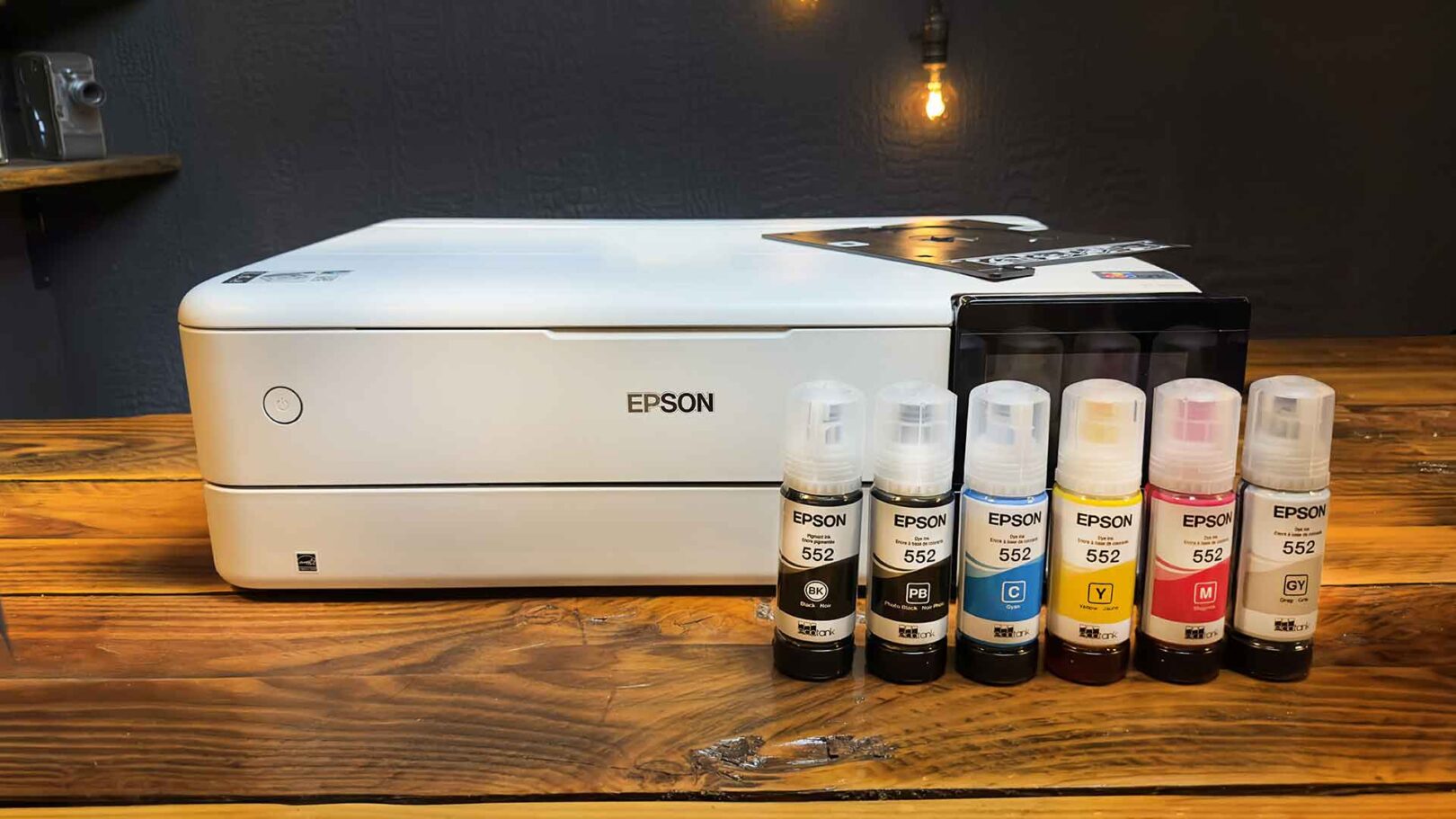 A white Epson Ecotank ET-8550 printer sits on a wooden table next to six Epson 552 ink bottles in black, cyan, magenta, yellow, gray, and photo black—perfect for fueling your creativity.