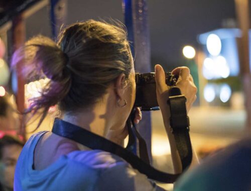 Artist who travels with brown hair in a ponytail taking a photo with a camera at night, city lights blurred in the background.