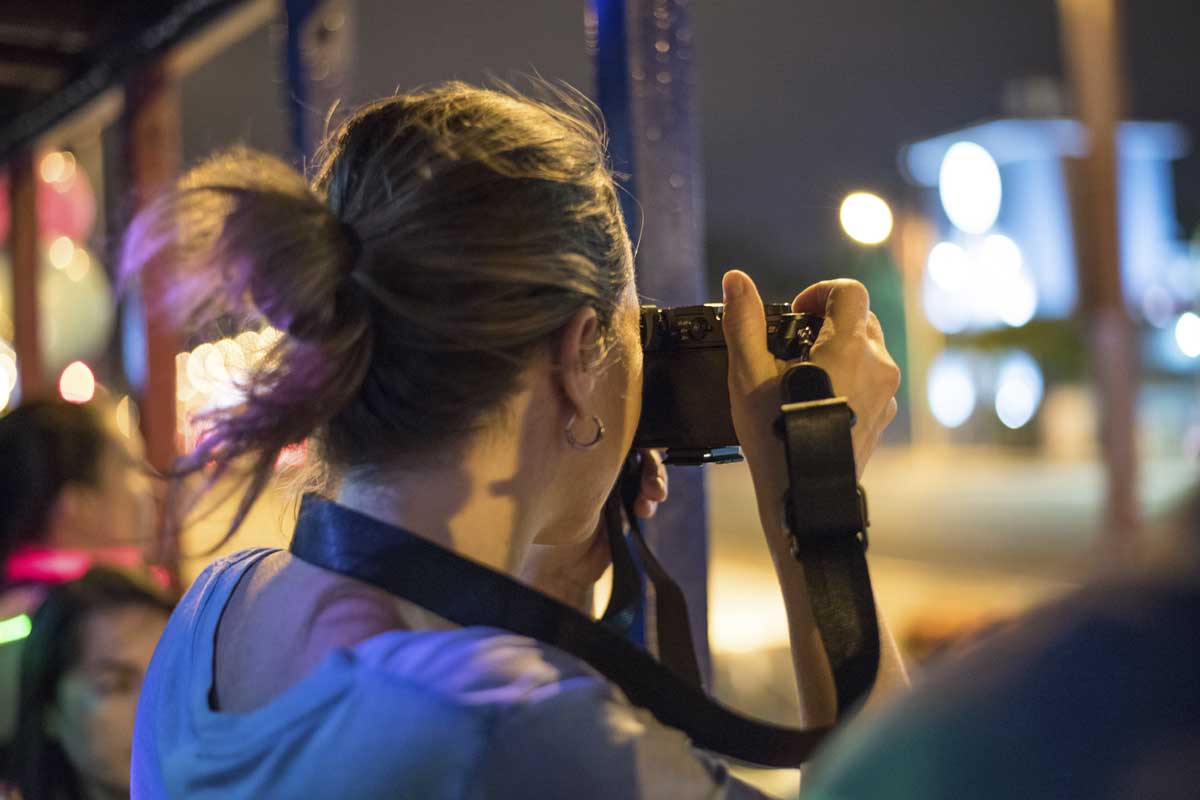 Artist who travels with brown hair in a ponytail taking a photo with a camera at night, city lights blurred in the background.