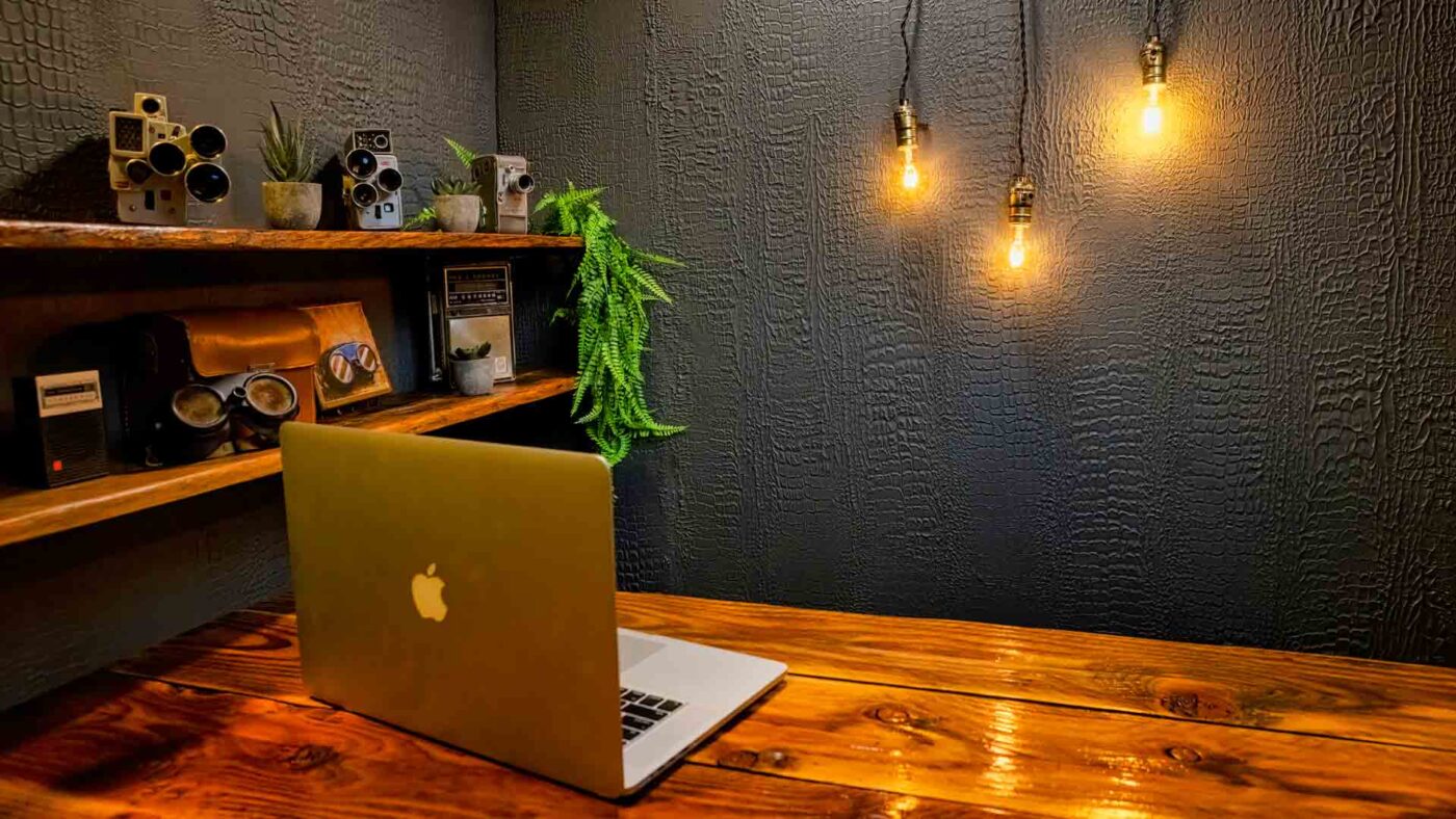 A wooden desk with an open MacBook, shelf with retro cameras and plants, and three exposed hanging light bulbs against a textured dark wall—perfect for a stylish DIY YouTube Studio or cozy home video studio setup.