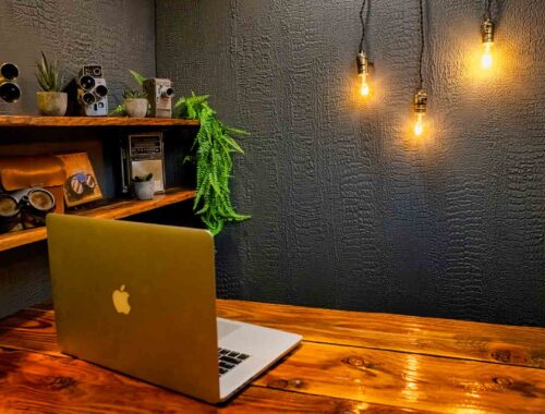 A wooden desk with an open MacBook, shelf with retro cameras and plants, and three exposed hanging light bulbs against a textured dark wall—perfect for a stylish DIY YouTube Studio or cozy home video studio setup.
