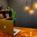 A wooden desk with an open MacBook, shelf with retro cameras and plants, and three exposed hanging light bulbs against a textured dark wall—perfect for a stylish DIY YouTube Studio or cozy home video studio setup.