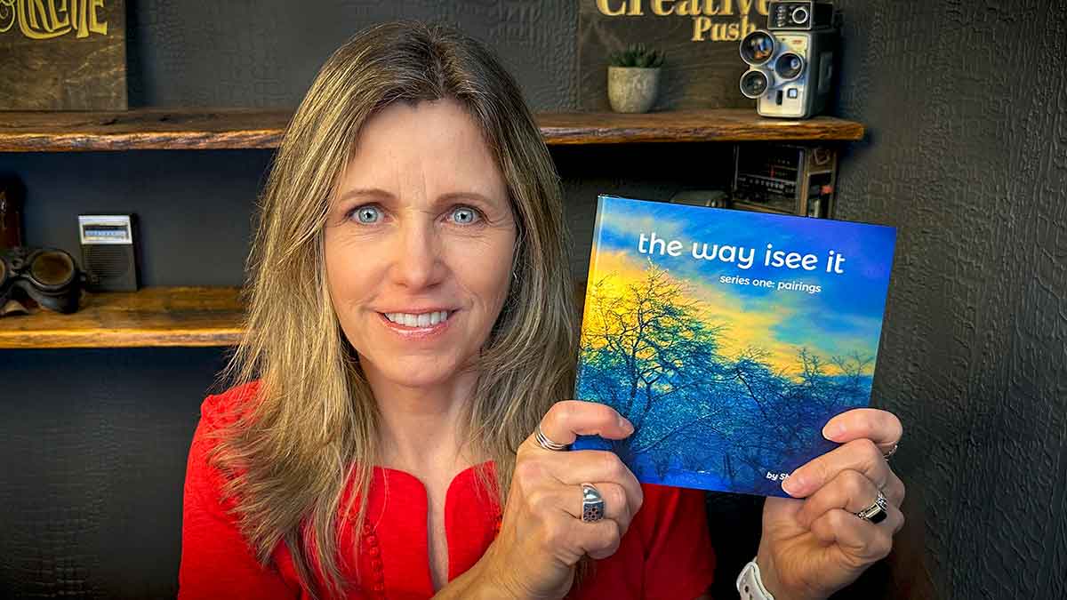 A woman in a red top holds up her Blurb print-on-demand book, "the way i see it: series one, pairings," in front of a shelf with cameras and books.